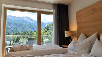 Habitación de hotel con cama, mesita de noche, lámpara y vista a montañas a través de un ventanal grande.