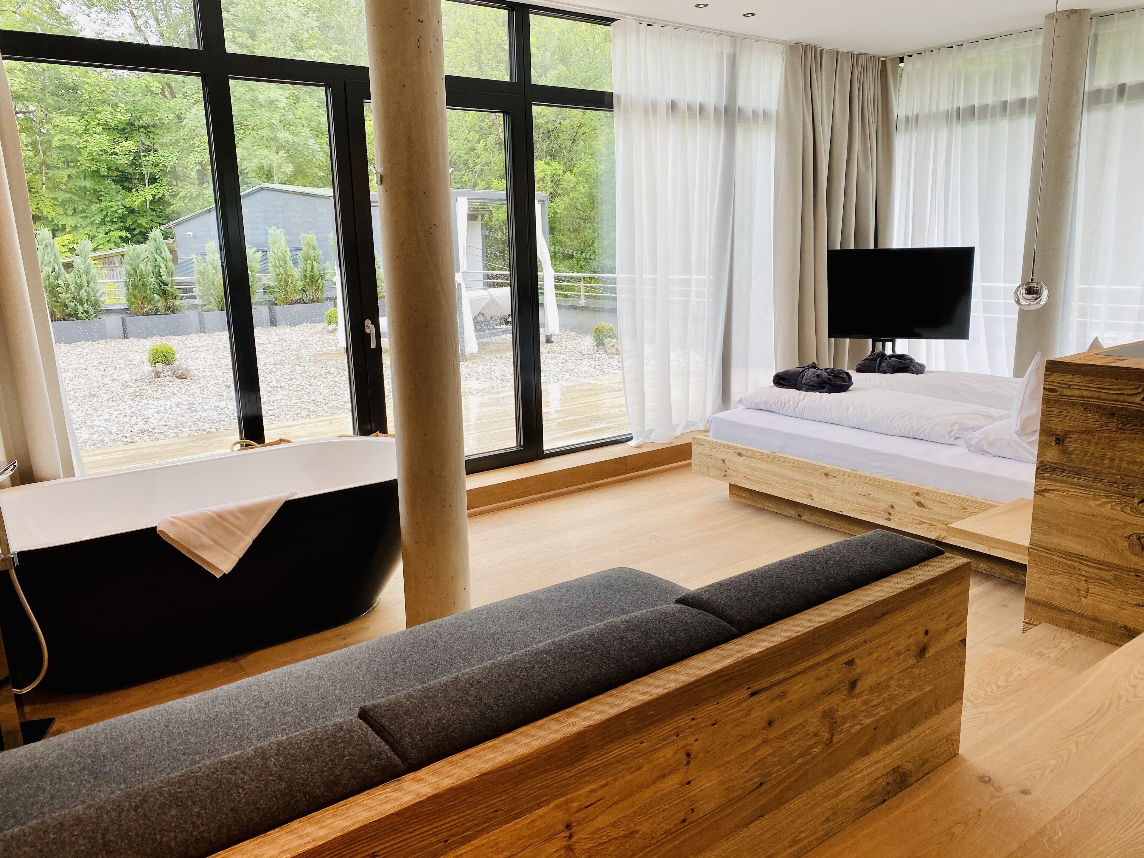 A modern bedroom with a large window, a wooden bed, a TV, and a bathtub.