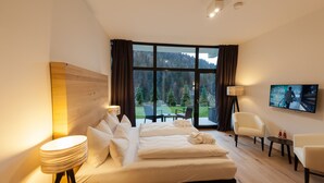 Een hotelkamer met een groot bed, twee nachtkastjes, een televisie en uitzicht op een bos door grote ramen.