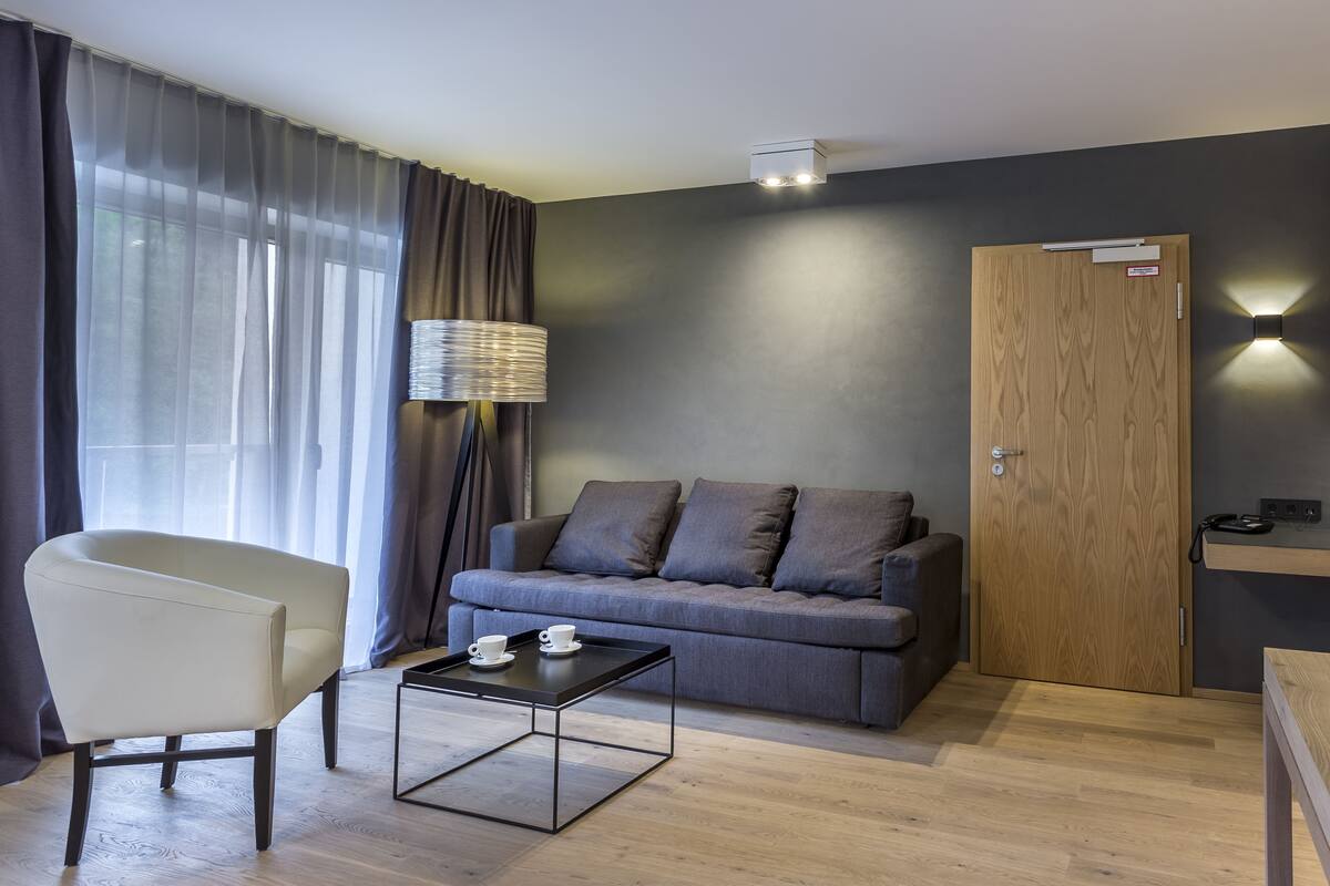 Een moderne kamer met een grijze bank, een witte fauteuil, een klein tafeltje met kopjes, een lamp en een houten deur.