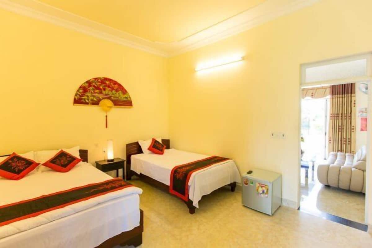 A hotel room with two single beds, a small table, a nightstand, a lamp, a fan, and a decorative wall hanging.