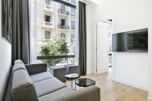 A modern living room with a grey sofa, two armchairs, a small round table, and a TV on the wall. - Murmuri Residence Mercader (Barcelona)