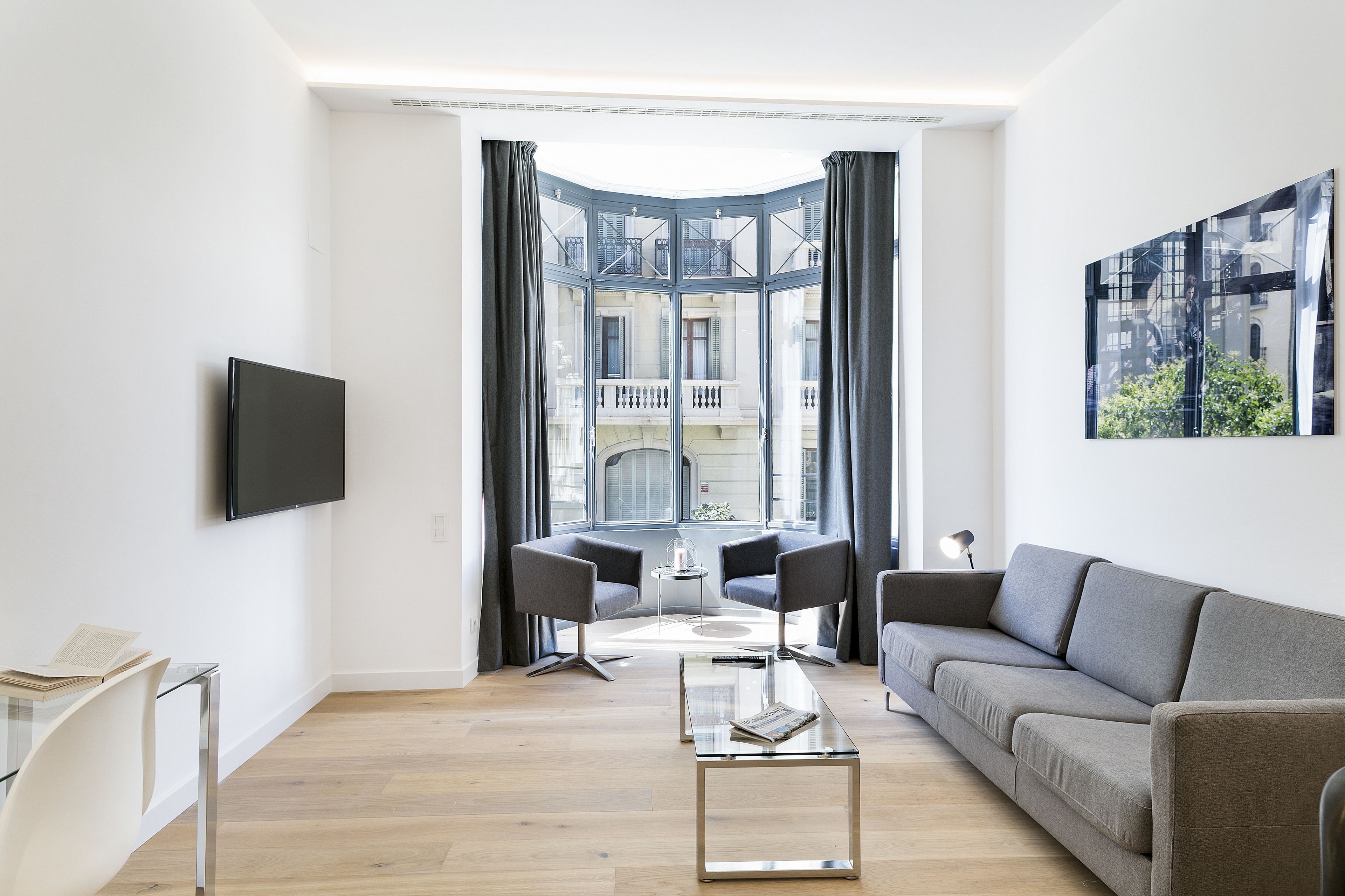 Ein modernes Wohnzimmer mit einem grauen Sofa, einem Glastisch und zwei Sesseln. Ein Flachbildfernseher ist an der Wand befestigt und ein großes Fenster bietet Blick auf die Stadt.