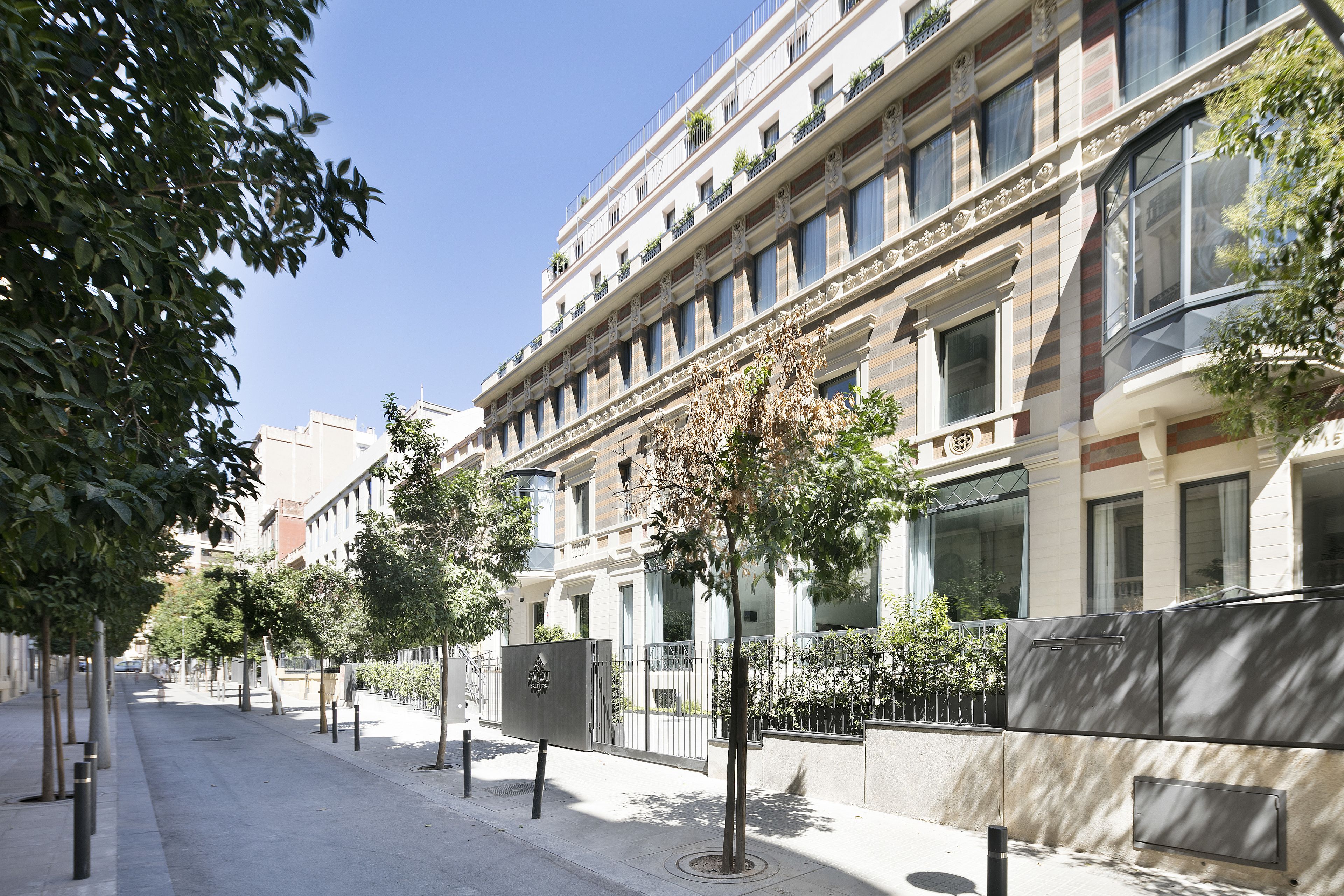 Vista de la calle de un edificio de varios pisos, con balcones y una acera bordeada de árboles.