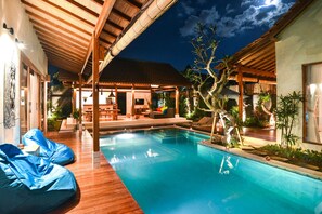 A swimming pool with wooden decking, a covered seating area, and a well-lit outdoor space with plants.