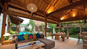 Un área de estar al aire libre con techo de madera, piscina al fondo y un conjunto de sofás y mesas.