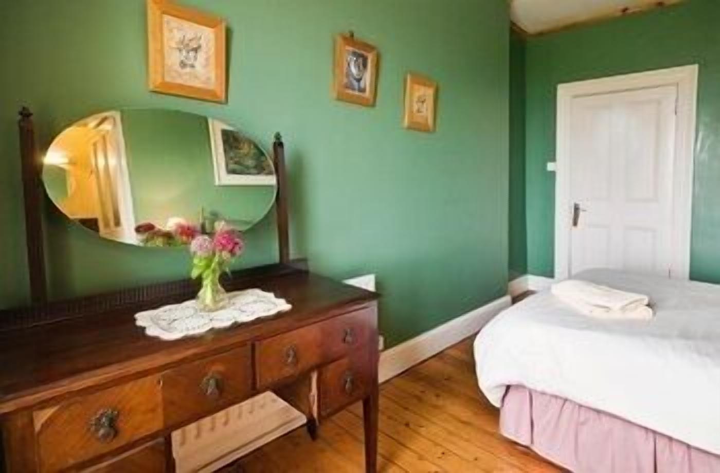 A bedroom with a wooden dresser, a mirror, a bed with a white comforter, and green walls.
