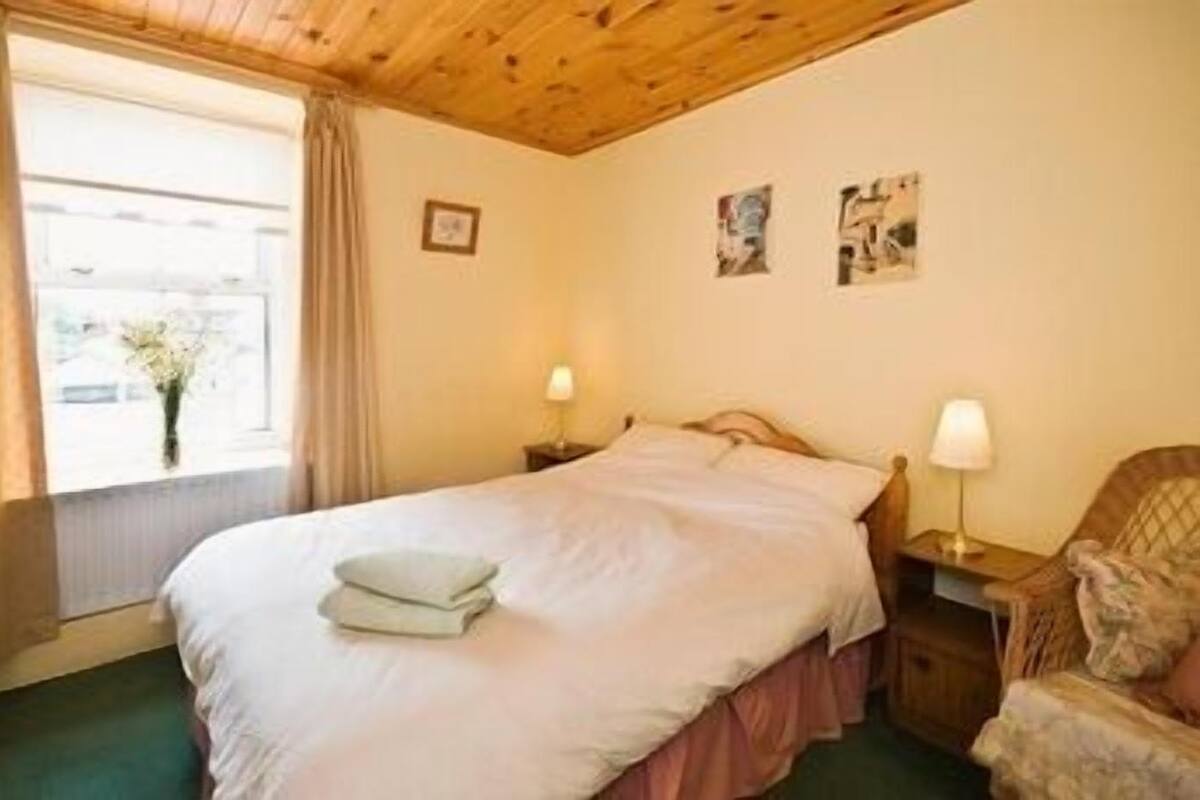 A bedroom with a bed, two bedside lamps, a window with curtains, and a chair.