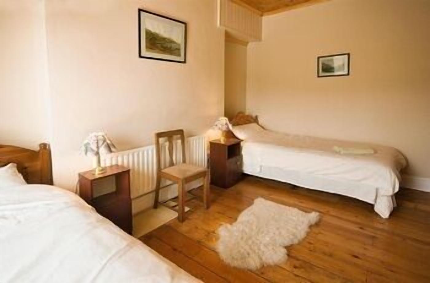 A room with two single beds, a wooden floor, a radiator, a chair, and a small table with a lamp.