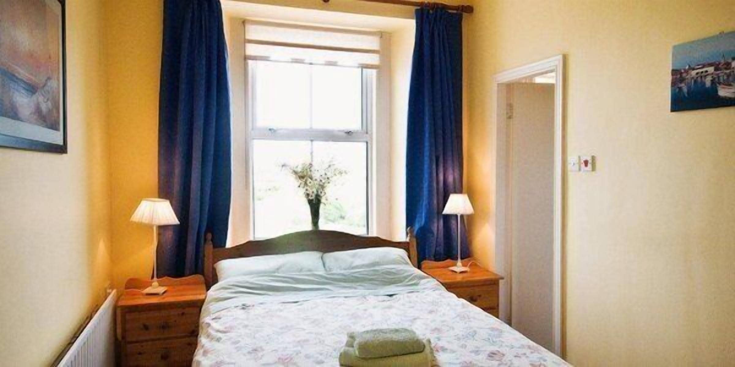 A bedroom with a bed, two bedside tables, a window with curtains, a door, and a wall-mounted picture.