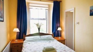 A bedroom with a bed, two bedside tables, a window with curtains, a door, and a wall-mounted picture.