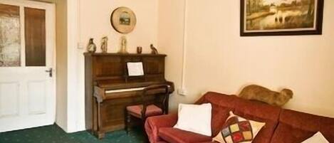 Een kamer met een houten piano, een rode bank met kussens, een ingelijst schilderij aan de muur en een deur.