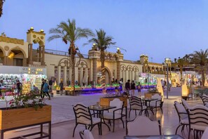 An outdoor dining area with tables and chairs, palm trees, and a building with arches and a sign that reads "SOLO LAMA AICA BEVER". - Cataract Layalina Resort (Sharm El Sheikh)