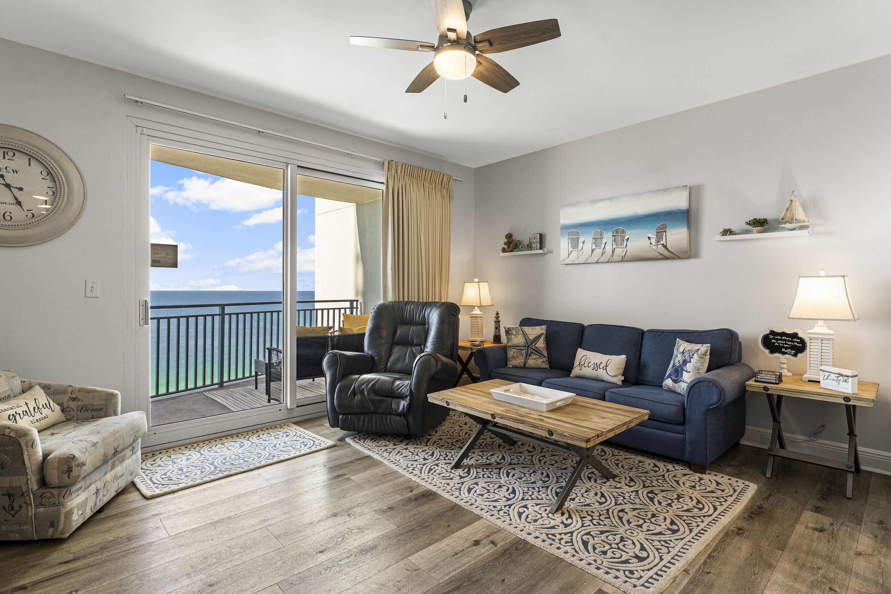 A living room with a view to the balcony, a ceiling fan, a clock, and a sofa with a patterned rug.