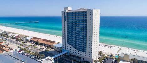 A high-rise hotel with a beach view, parking lots, and a pier in the background.