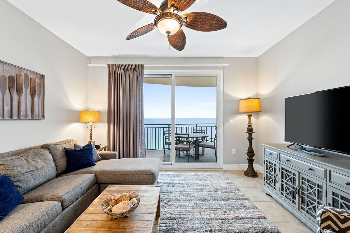 Ein modernes Wohnzimmer mit Deckenventilator, einer Couch, einem Couchtisch, einem Fernseher und einem Balkon mit Blick.