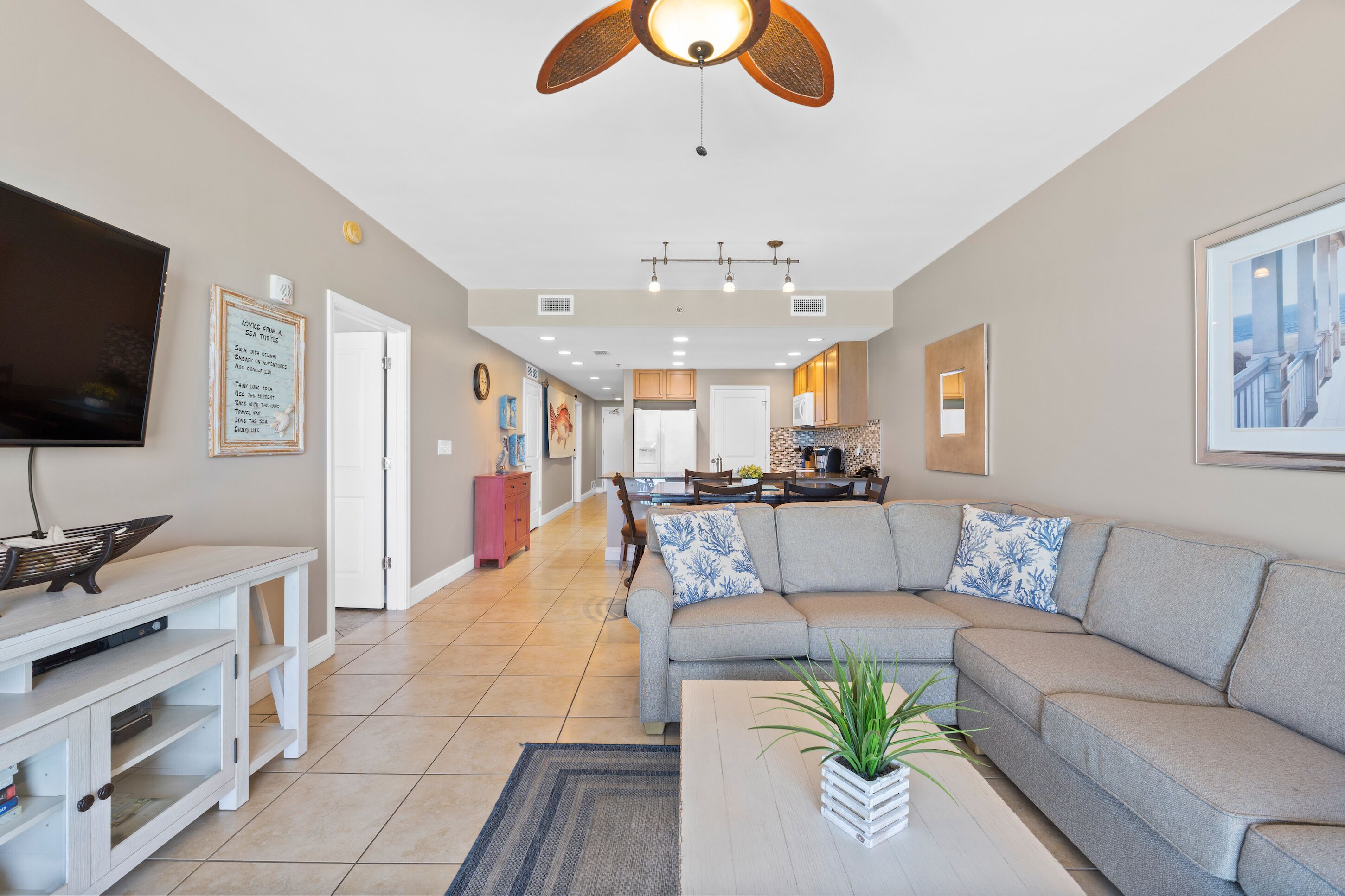 A spacious living room with a large grey sectional sofa, a flat-screen TV, and a dining area with a kitchen in the background.