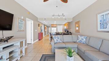 A spacious living room with a large grey sectional sofa, a flat-screen TV, and a dining area with a kitchen in the background.