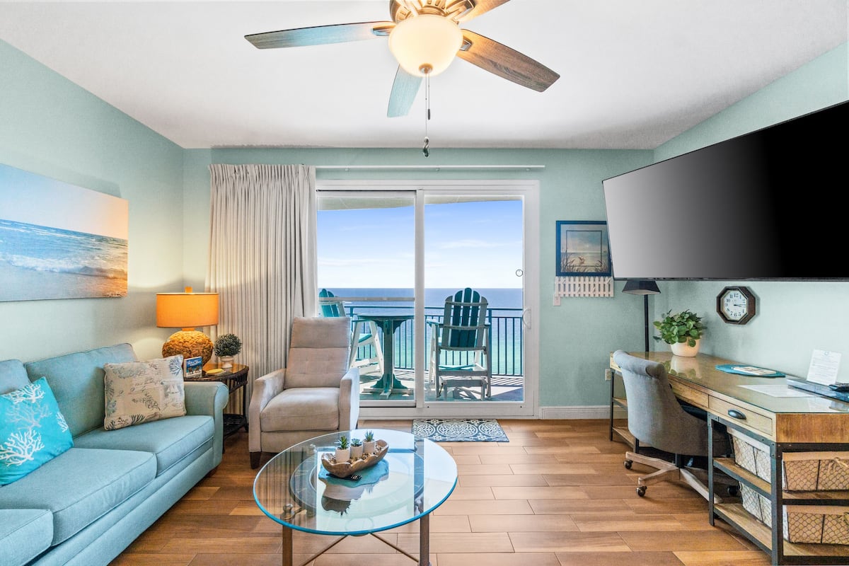 Ein Wohnzimmer mit Sofa, Sessel, Glastisch und Schreibtisch mit Computer. Ein Fernseher ist an der Wand montiert, ein Deckenventilator sorgt für Luftzirkulation und durch eine Glastür bietet sich Blick auf das Meer.