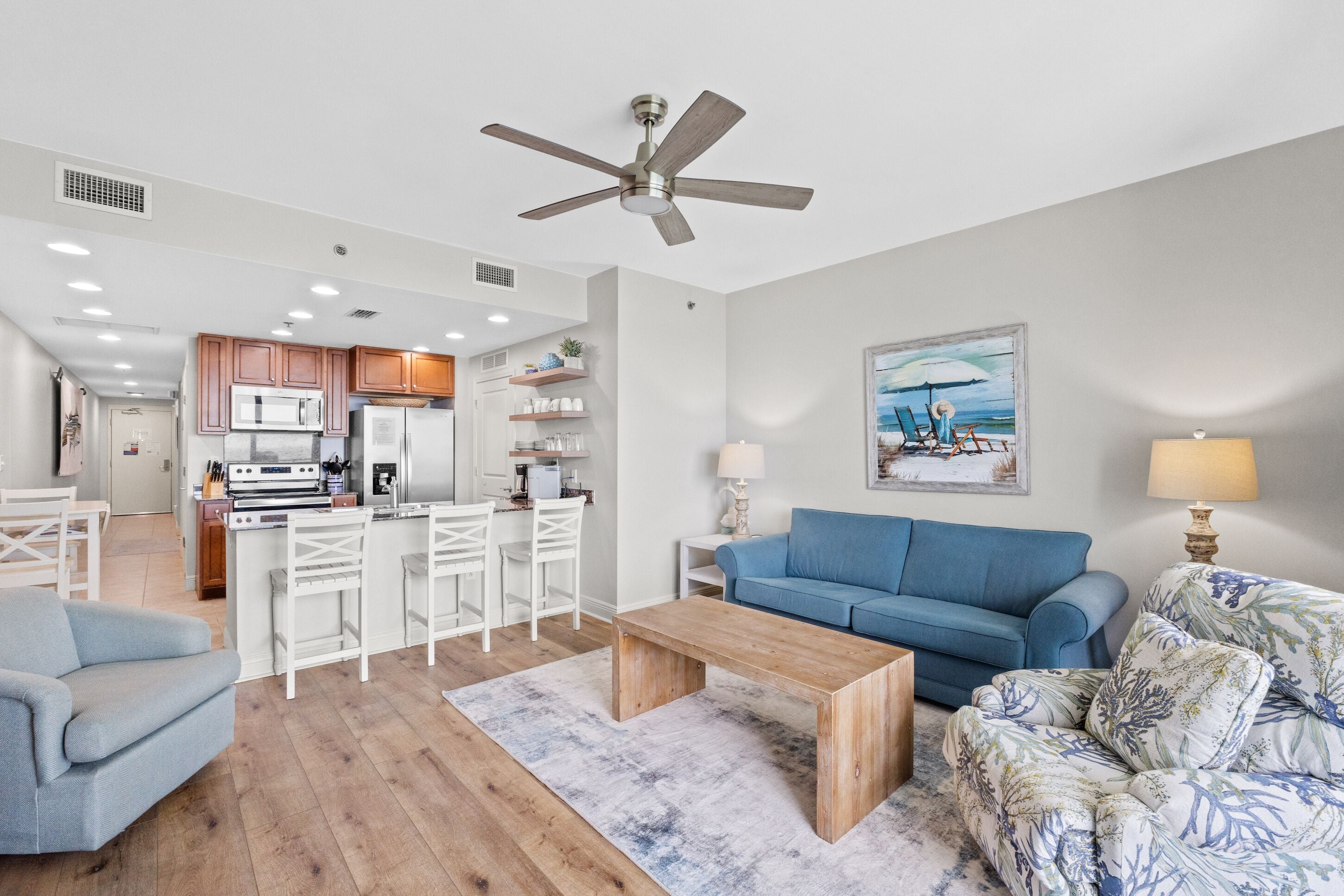 A modern living room with a blue sofa, a wooden coffee table, and a patterned armchair. There is a kitchen area with white cabinets and a microwave.