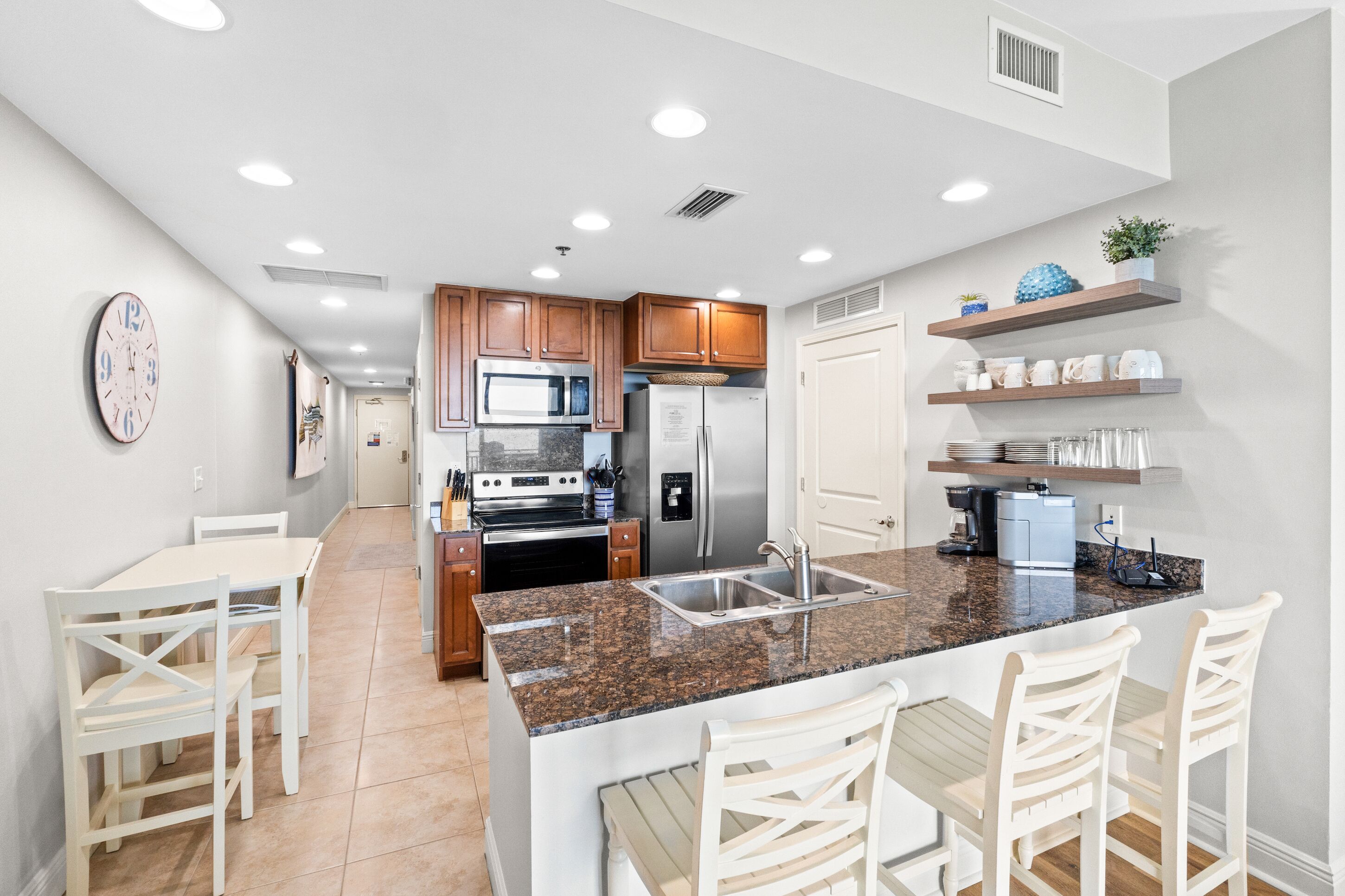 A modern kitchen with granite countertops, stainless steel appliances, and wooden cabinets. There is a dining area with white chairs and a table.