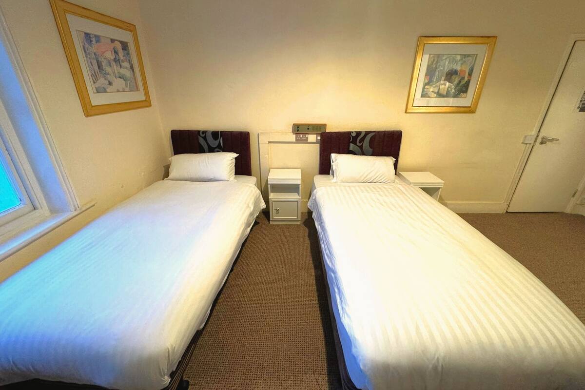 A hotel room with two single beds, a nightstand with a lamp, a small table, and a door.