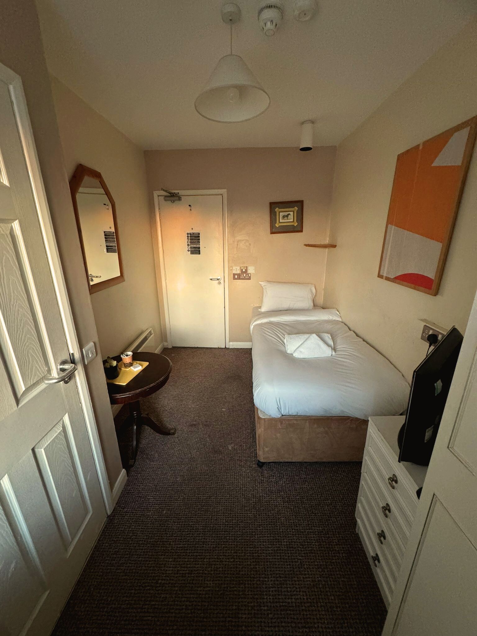 A hotel room with a bed, a desk with a chair, a dresser, a mirror, and a door.