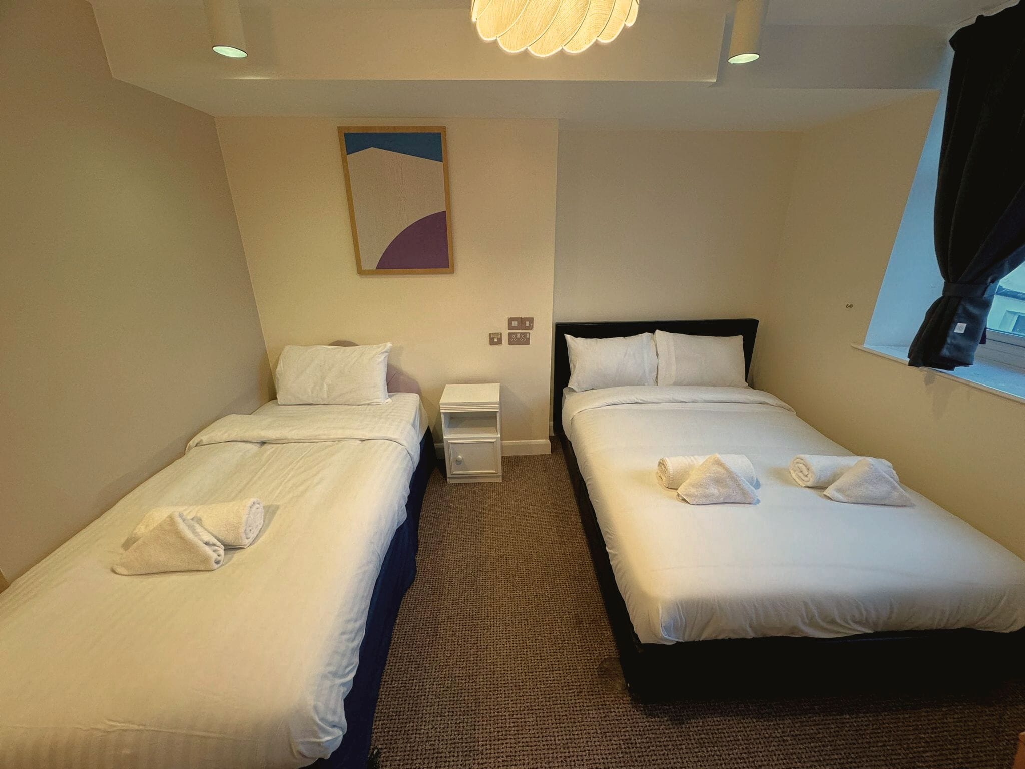 A hotel room with two single beds, a nightstand, a lamp, and a window with curtains.