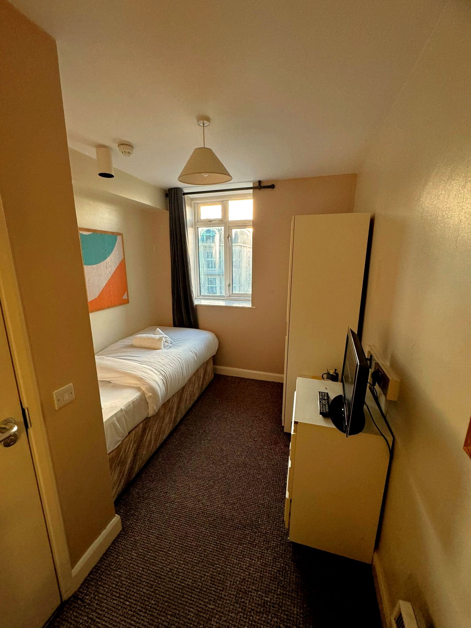 Ein kleines Hotelzimmer mit einem Einzelbett, einem kleinen Schreibtisch mit Computer, einem Fenster mit Vorhängen und einer Deckenleuchte.