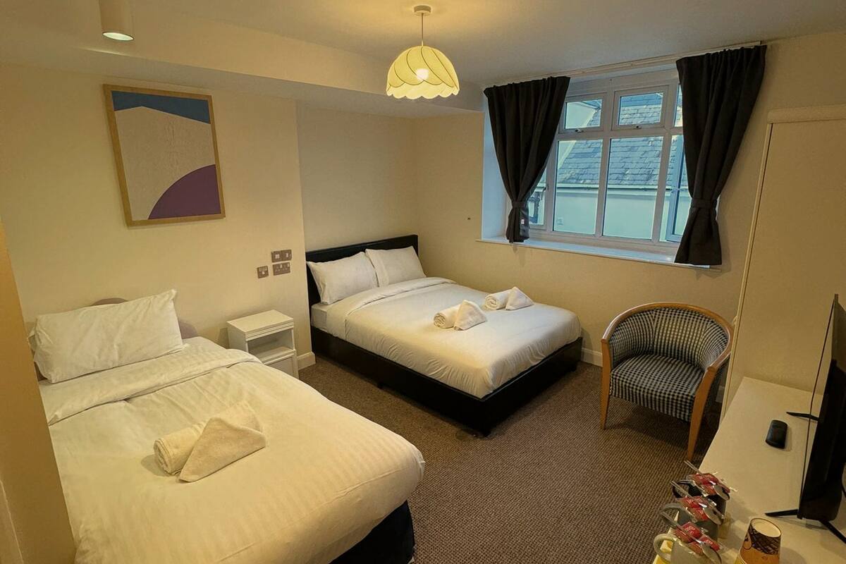A hotel room with two beds, a chair, a desk with a computer, a window with curtains, and a painting on the wall.