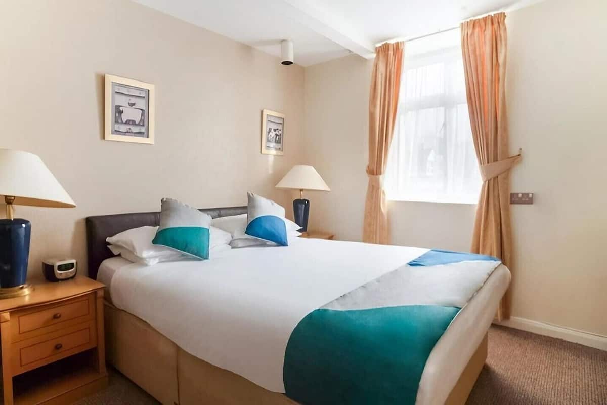 A hotel room with a bed, two bedside tables, two lamps, a nightstand, a window with curtains, and two framed pictures on the wall.