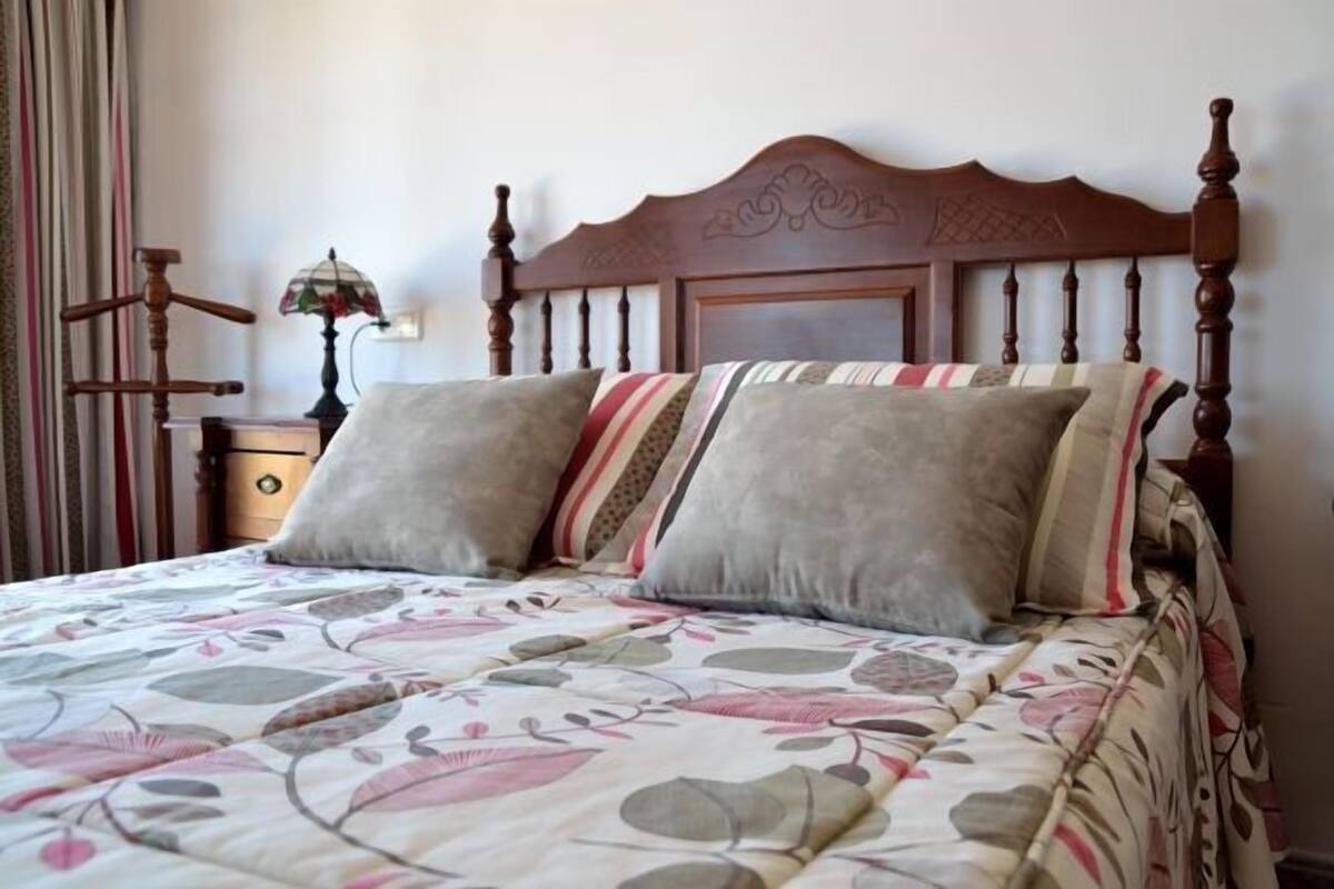 A bed with a floral-patterned bedspread and two pillows, a wooden headboard, a nightstand with a lamp, and a wall-mounted mirror.