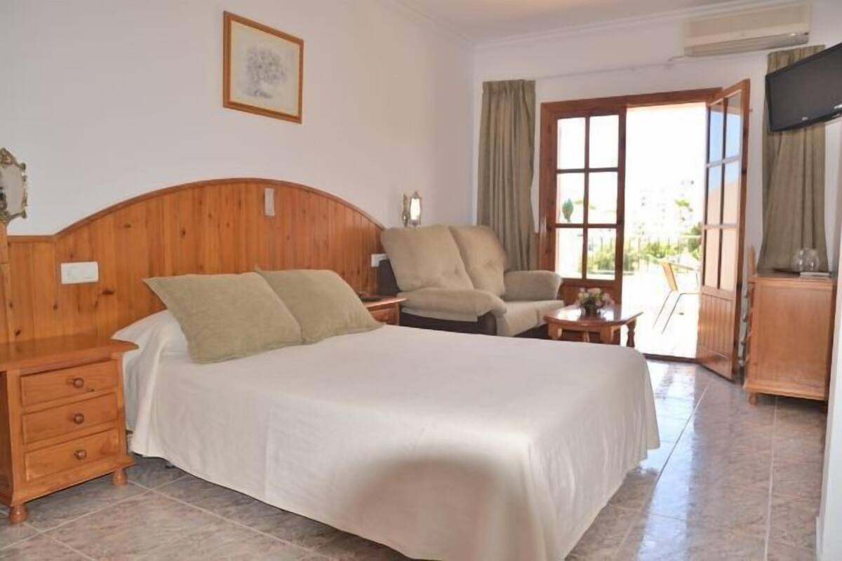 A bedroom with a wooden headboard bed, a wooden nightstand, a chair, a small table, and a TV on a stand.