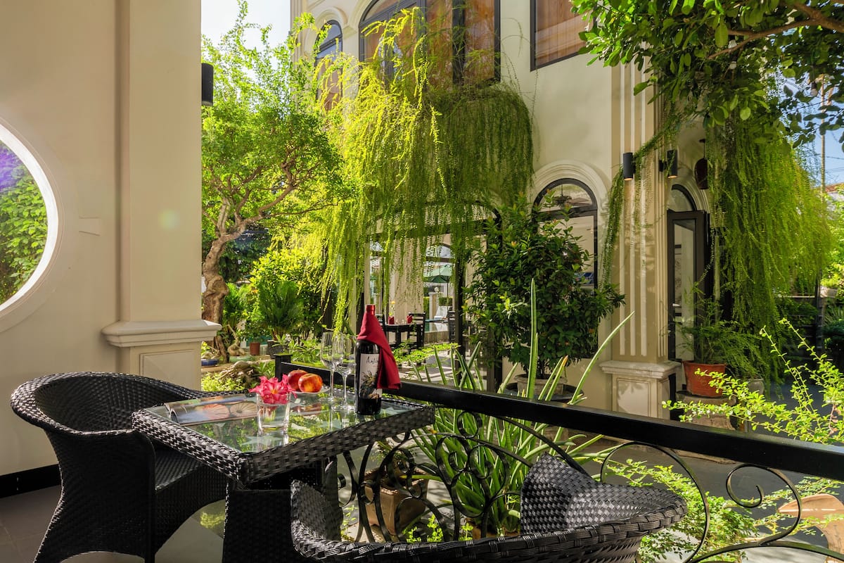 A balcony with wicker chairs and a glass table, surrounded by lush greenery and a building with arched windows.