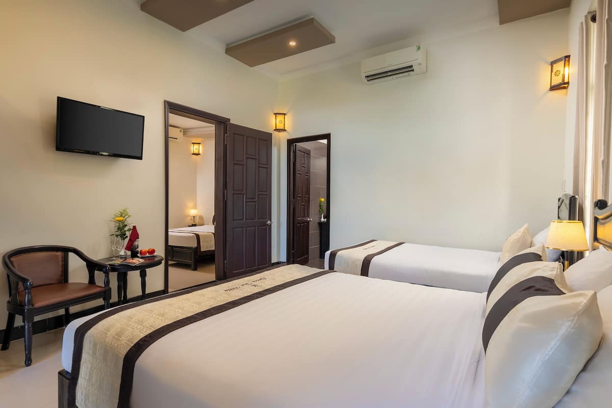 A hotel room with two beds, a TV, a desk with a chair, and a small table with a vase and fruit.