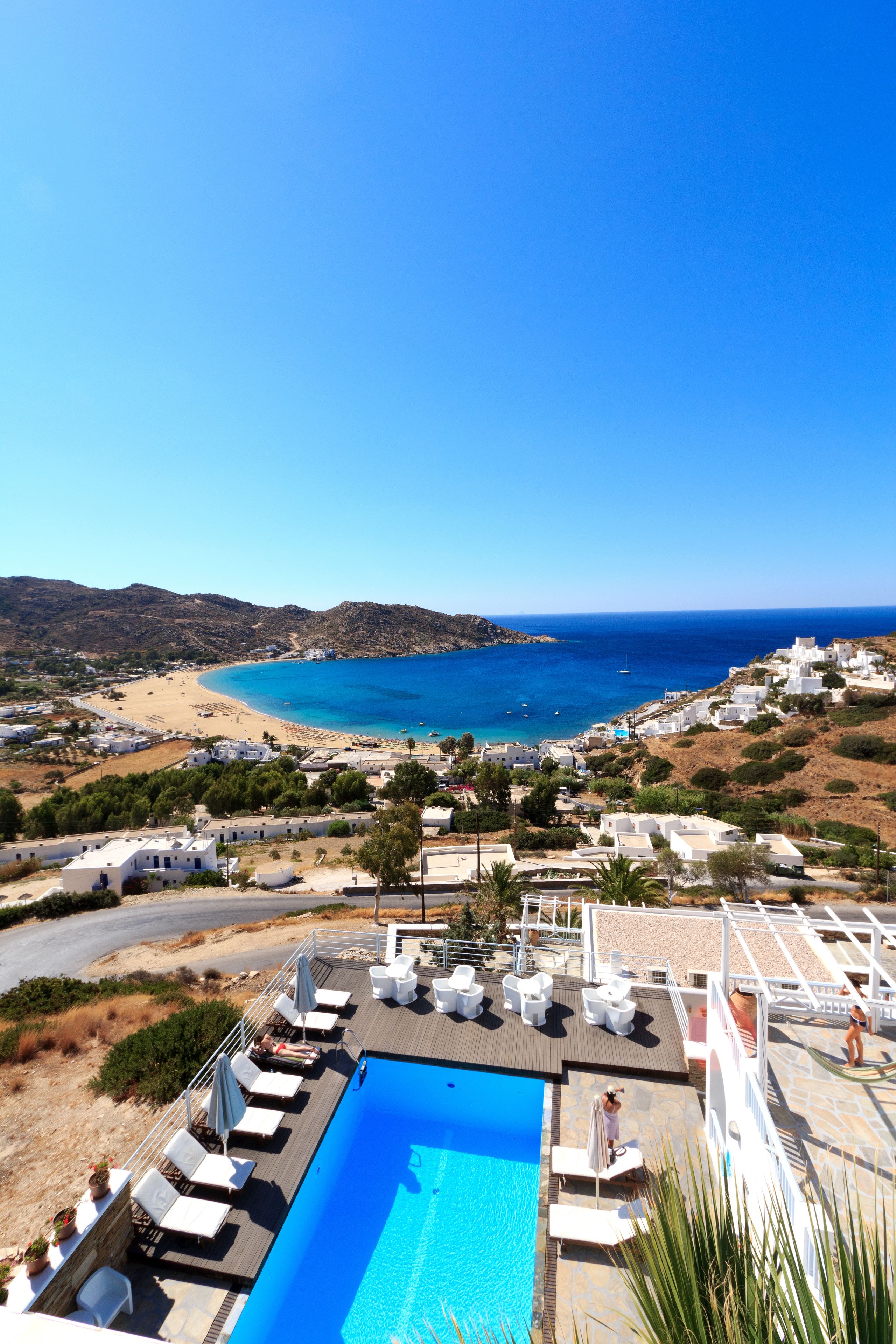 A pool area with lounge chairs, a clear view of the sea, and a mountainous landscape in the background.