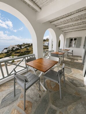 A covered outdoor seating area with stone flooring, white walls, and a view of a coastal village. - Far Out Hotel & Spa (Ios)