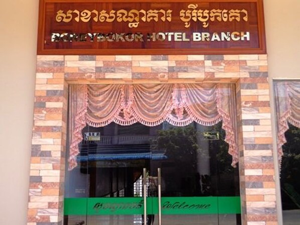 A hotel entrance with a sign in a local language and English, a brick facade, and a green sign with the word "Welcome".