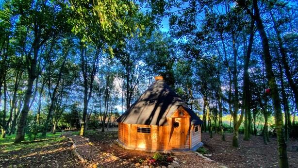 A wooden cabin in a forest with a thatched roof, surrounded by trees and a dirt path. - High Parks (Bedale)
