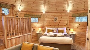 A cozy wooden cabin room with a bed, two bedside tables with lamps, a sofa with cushions, and a window with a view of trees. - High Parks (Bedale)