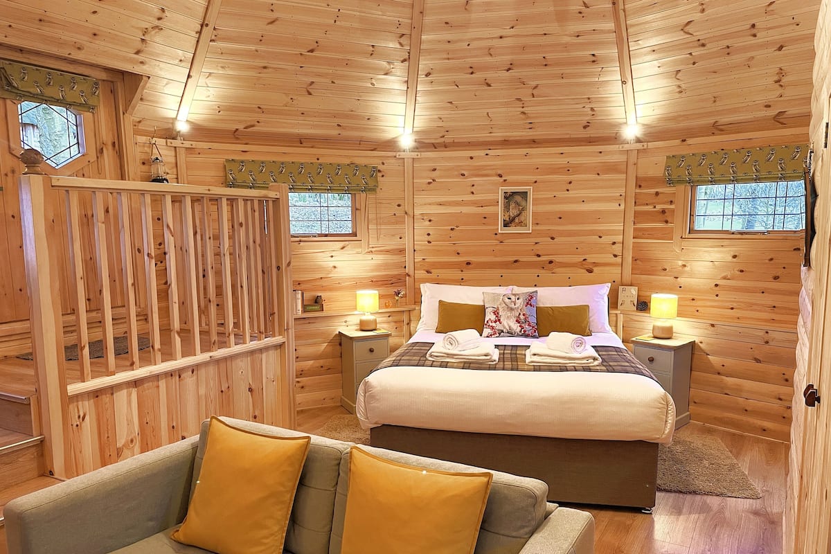 A cozy wooden cabin room with a bed, two bedside tables with lamps, a sofa with cushions, and a window with a view of trees.