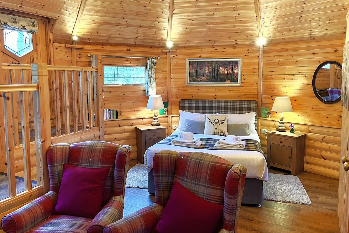A cozy log cabin room with a plaid bedspread, two plaid armchairs, a wooden ceiling, and a framed picture on the wall.