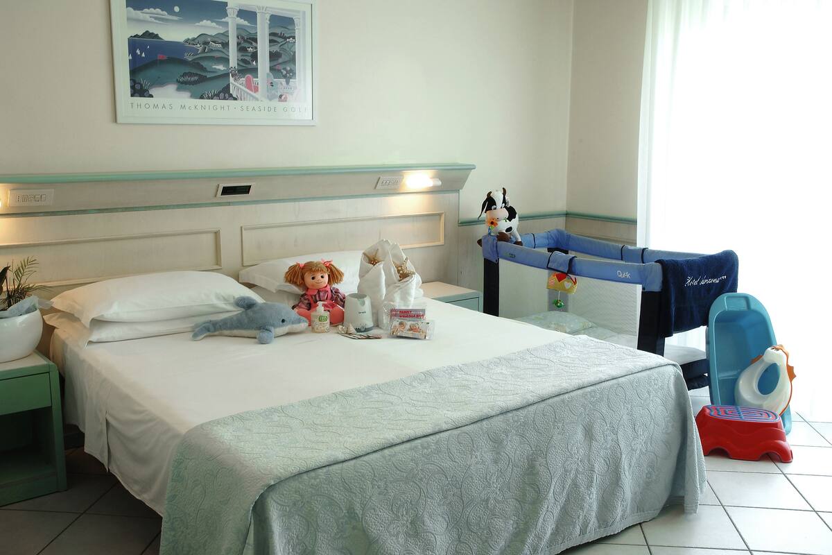 A hotel room with a bed, pillows, stuffed toys, a nightstand, a wall-mounted clock, and a window with curtains.