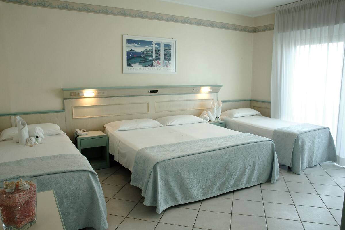 A hotel room with two single beds, a double bed, a bedside table with a lamp, a phone, and a painting on the wall.