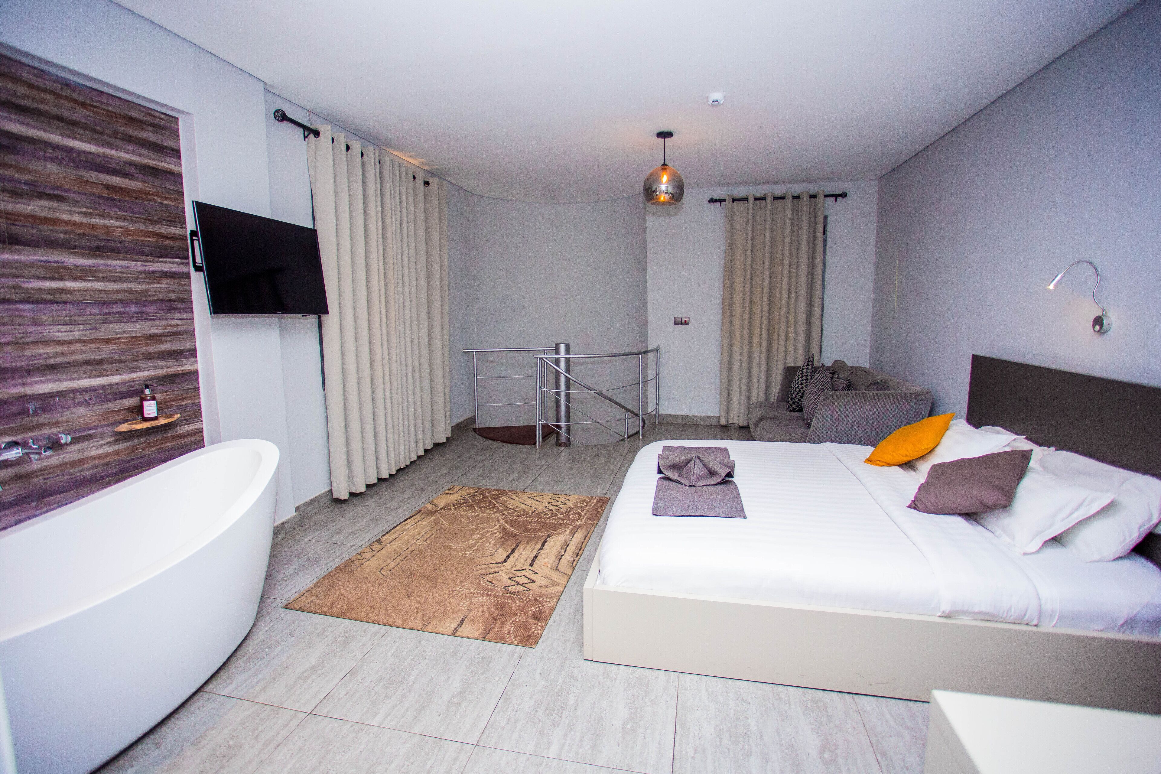 Una habitación de hotel moderna con una cama grande, un baño independiente con bañera, un televisor montado en la pared y una lámpara colgante.