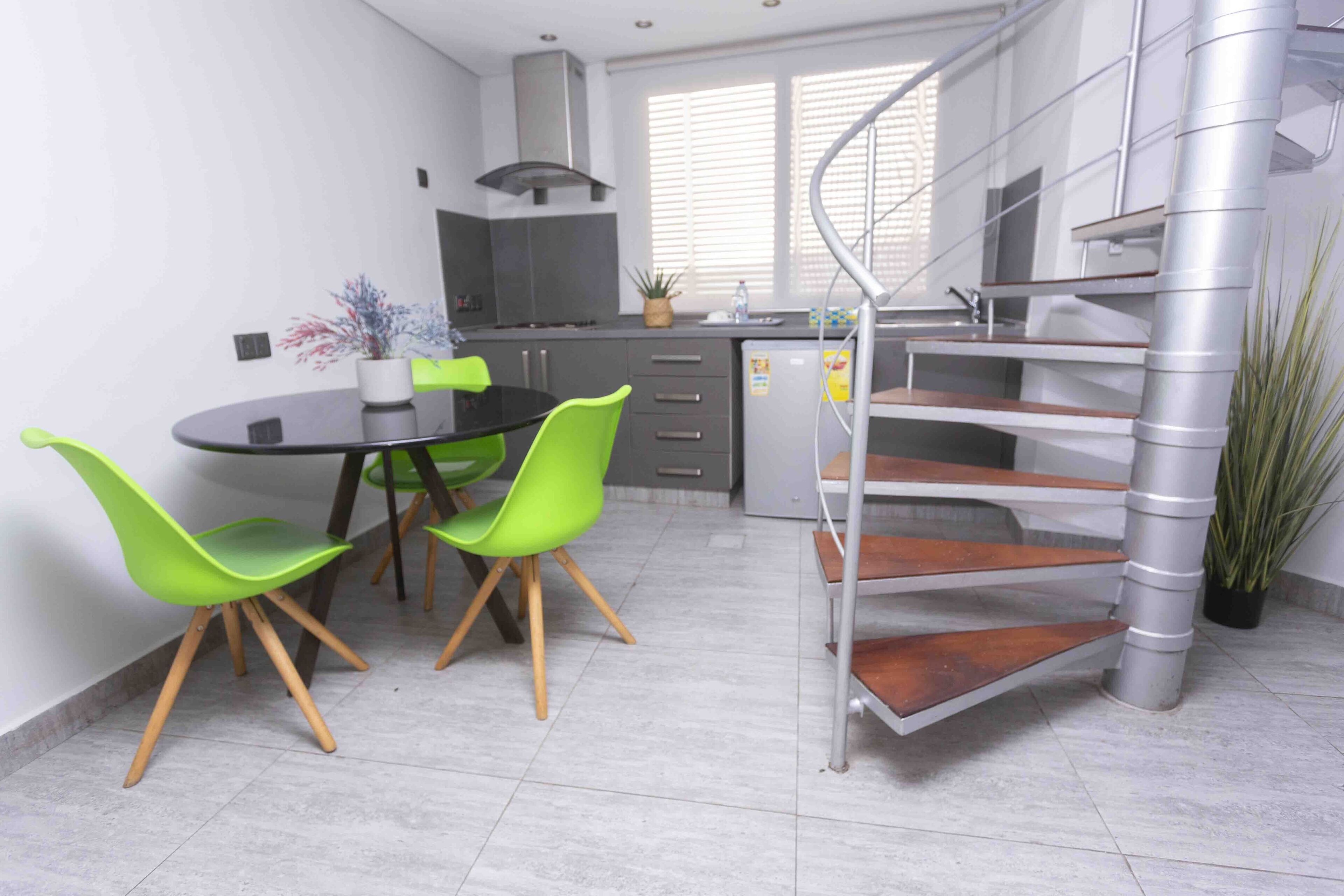 A modern kitchen with a dining area, featuring a spiral staircase, grey cabinets, and a small potted plant.
