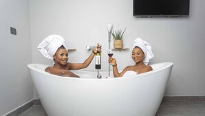 Two people in a modern, white bathtub with a wine bottle and glass, in a room with a flat-screen TV and a small plant on the wall. - Urbano Hotel (Accra)