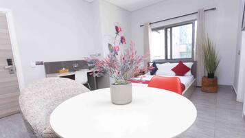 Habitación de hotel moderna con una mesa redonda blanca, un sofá estampado, una cama con almohadas rojas y blancas, una ventana con cortinas y una planta en maceta.