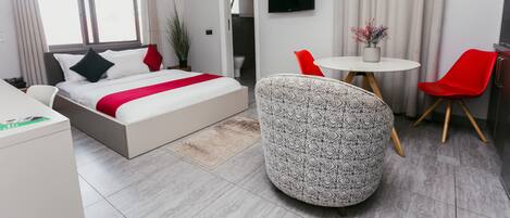 A modern hotel room with a bed, a desk, a round table with chairs, a TV, and a potted plant.