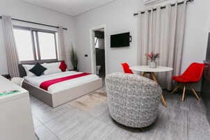 A modern hotel room with a bed, a desk, a round table with chairs, a TV, and a potted plant.